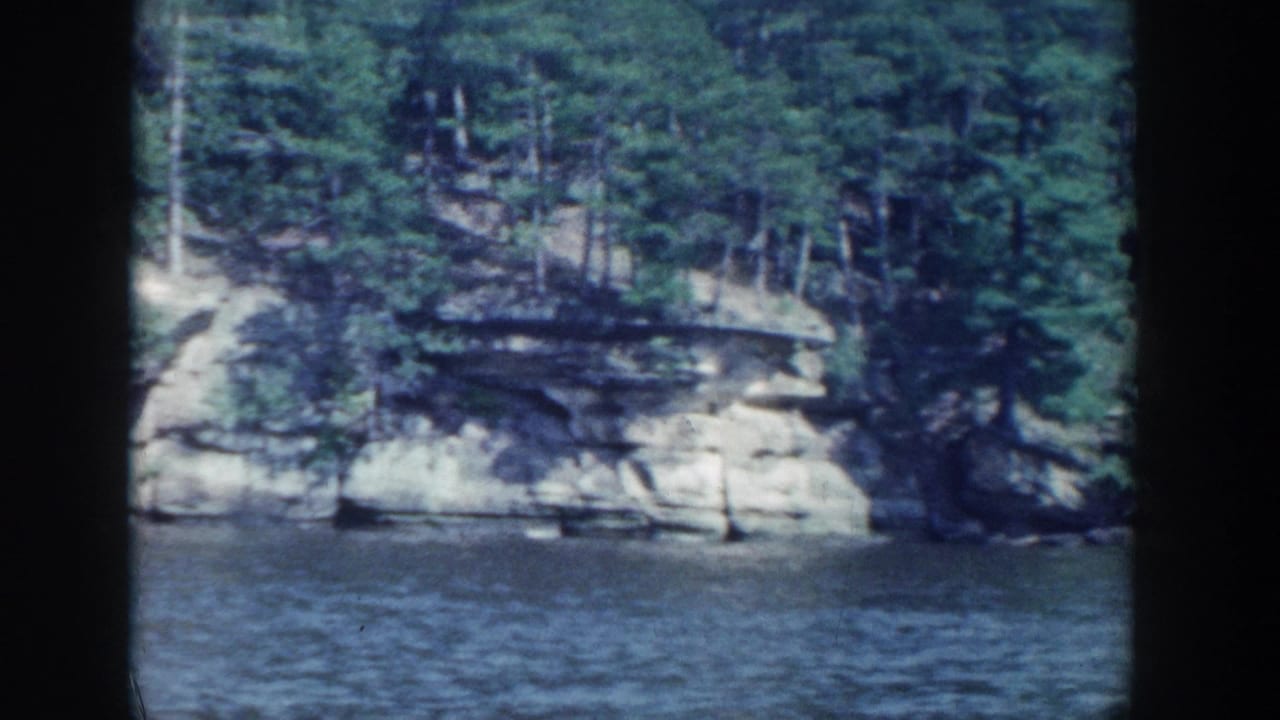 1964 Wisconsin 8mm frame showing a wooded rock ledge along calm water, bordered by the film gate.