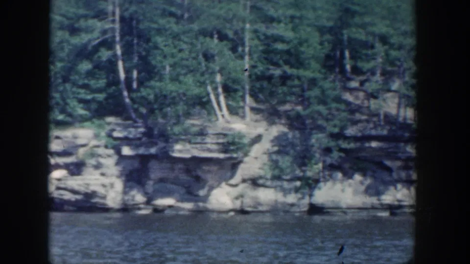 1964 Wisconsin 8mm frame with layered shoreline rock, dense trees, and still water in front.