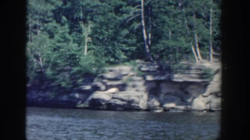 1964 Wisconsin home-movie frame showing a rocky wooded bank and calm water, with soft exposure and grain.