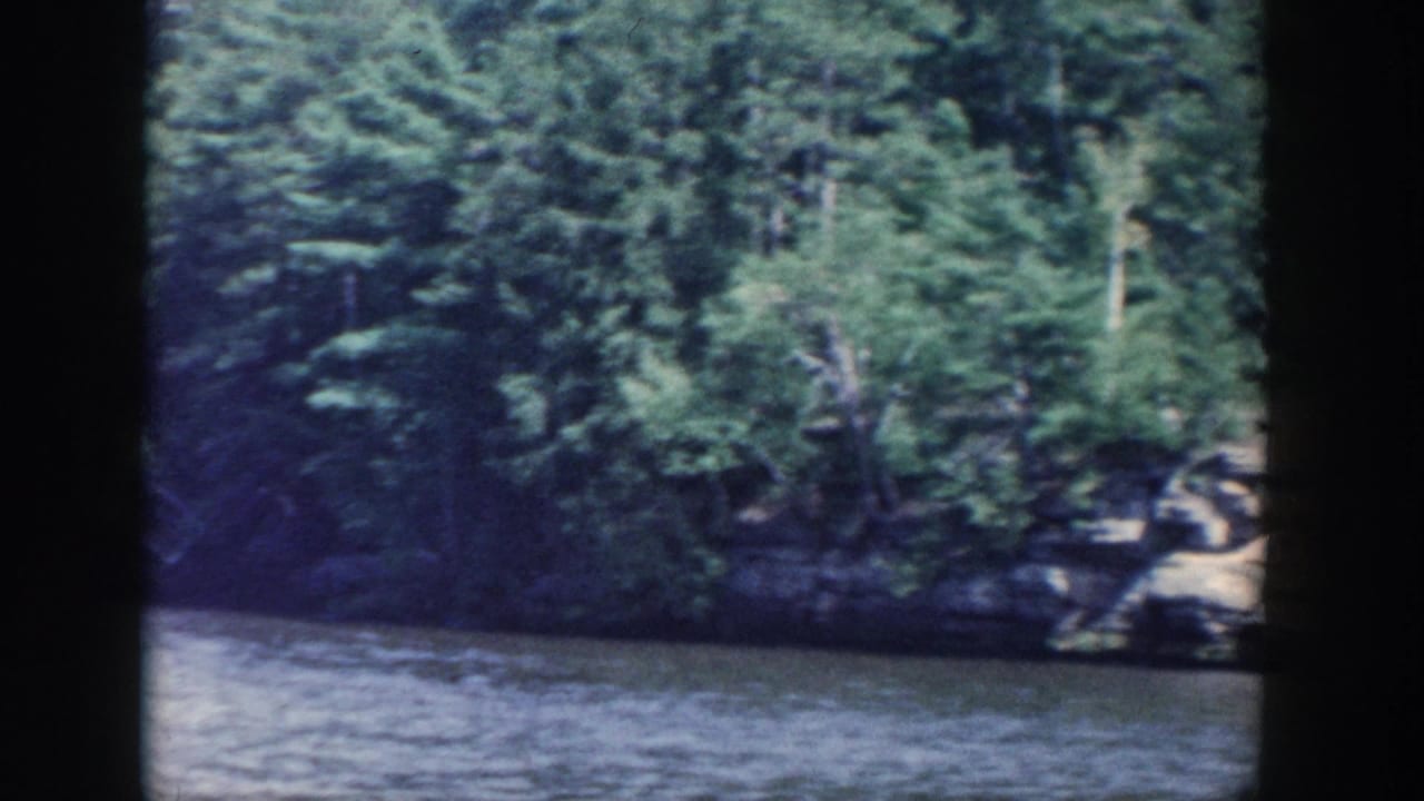 1964 Wisconsin 8mm frame showing a shaded wooded bank and water, with soft focus and visible film edge shadows.