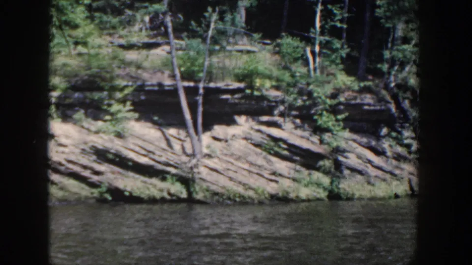 1964 Wisconsin 8mm frame showing a rocky, eroded shoreline with shrubs, thin tree trunks, and rippling water.