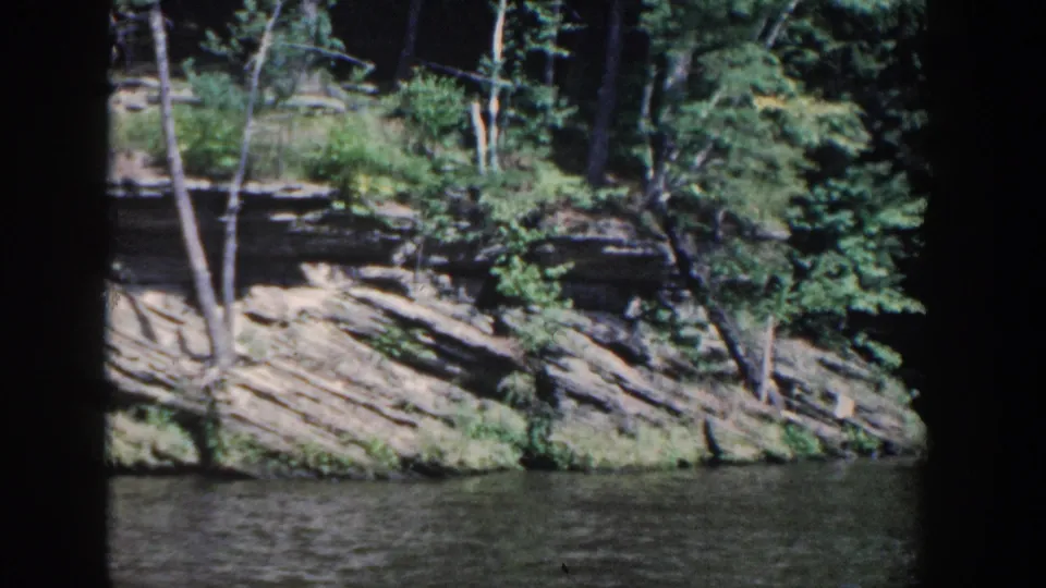 1964 Wisconsin 8mm frame of a forested shoreline with exposed sedimentary rock and dark home-movie frame edges.