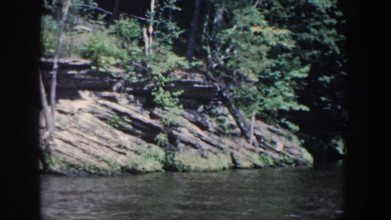 1964 Wisconsin 8mm view of a forested rock bank beside rippled river water, framed by the dark edge of a boat window.