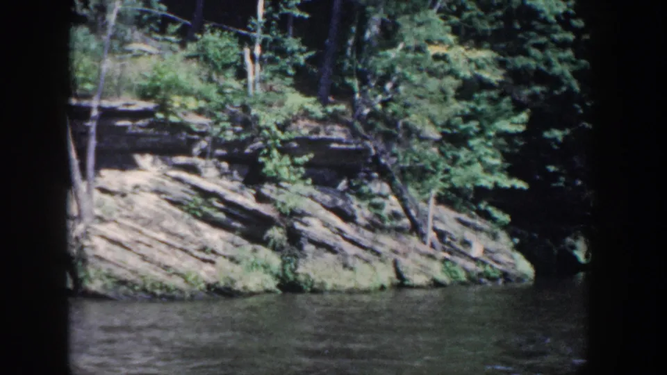 1964 Wisconsin 8mm view of a forested rock bank beside rippled river water, framed by the dark edge of a boat window.