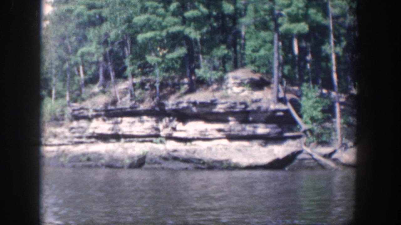 Forested Wisconsin riverbank with exposed horizontal rock strata and calm water, viewed from inside a boat in 8mm footage.