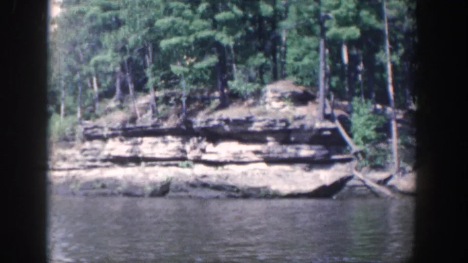 Forested Wisconsin riverbank with exposed horizontal rock strata and calm water, viewed from inside a boat in 8mm footage.