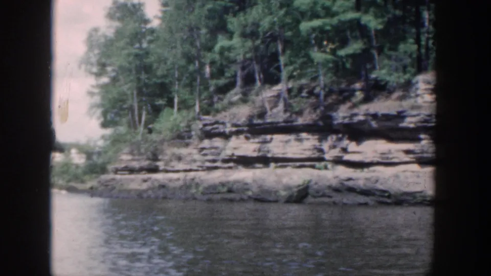 Vintage 8mm river view of a wooded Wisconsin shoreline with layered rock and water in the foreground.
