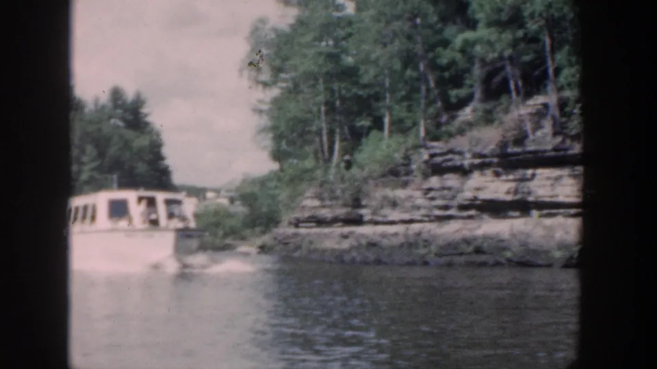 White motorboat with small visible passengers moves on a Wisconsin waterway beside trees and layered rock cliffs.
