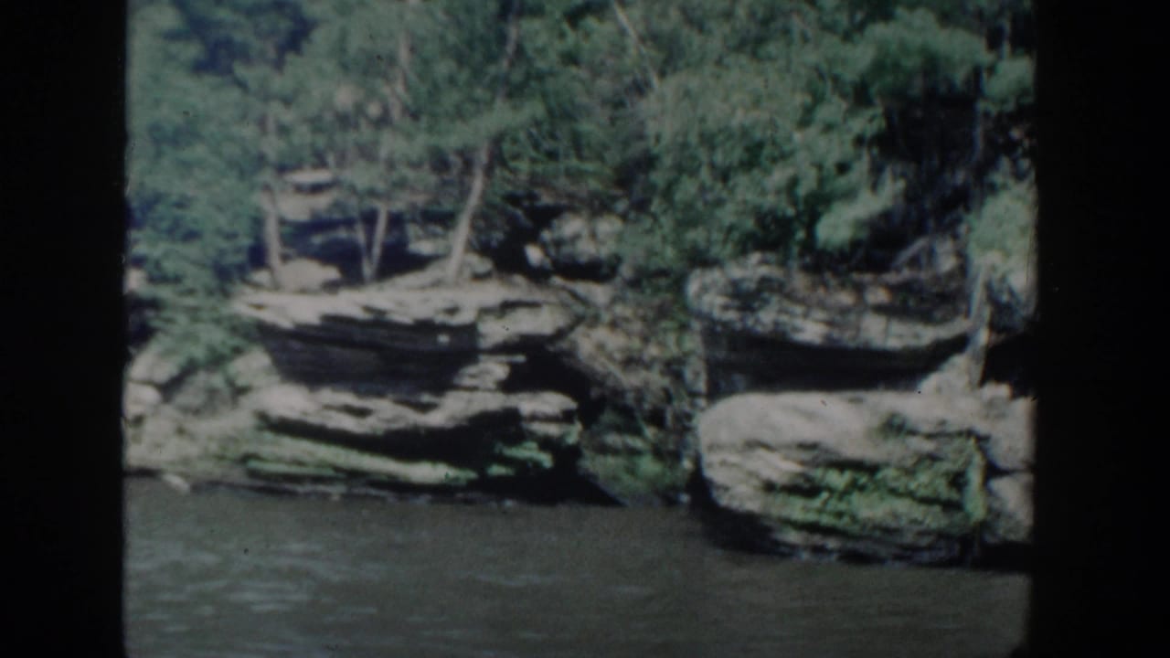 1964 Wisconsin 8mm frame showing layered riverbank cliffs, shrubs, and trees above calm rippled water.