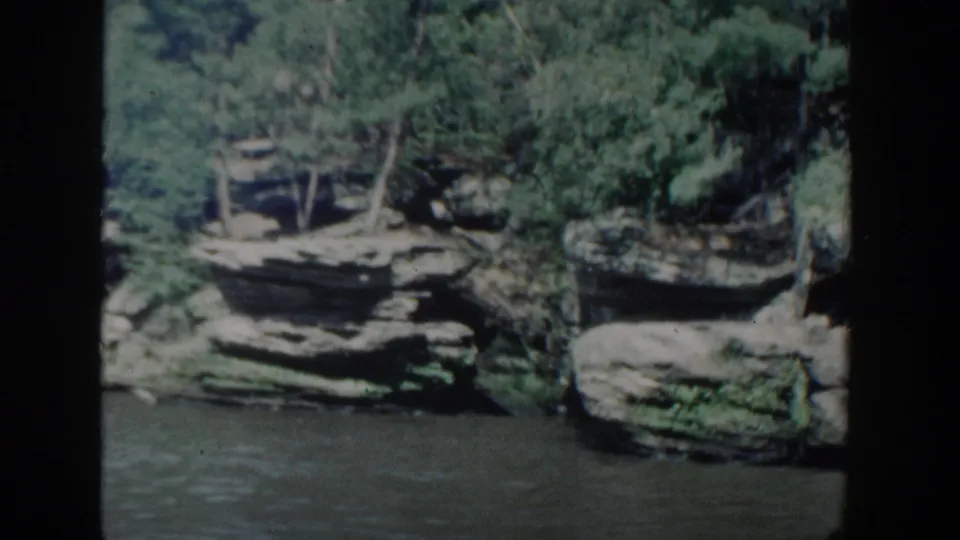 1964 Wisconsin 8mm frame showing layered riverbank cliffs, shrubs, and trees above calm rippled water.