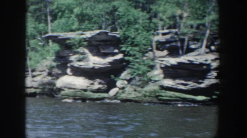 1964 Wisconsin 8mm frame showing sandstone-like ledges, trees, brush, and water from a moving river viewpoint.