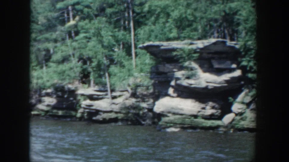 1964 Wisconsin 8mm frame showing a prominent rock overhang, river water, and dense green shoreline trees.
