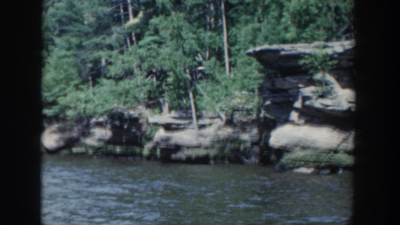1964 Wisconsin 8mm frame showing lake water, layered rock ledges, and dense trees through a dark window-like frame.