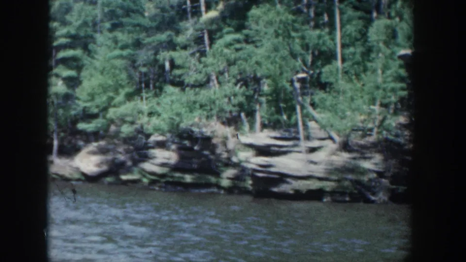 1964 Wisconsin shoreline view with rippled water, pale rock shelves, and dense trees along the bank.