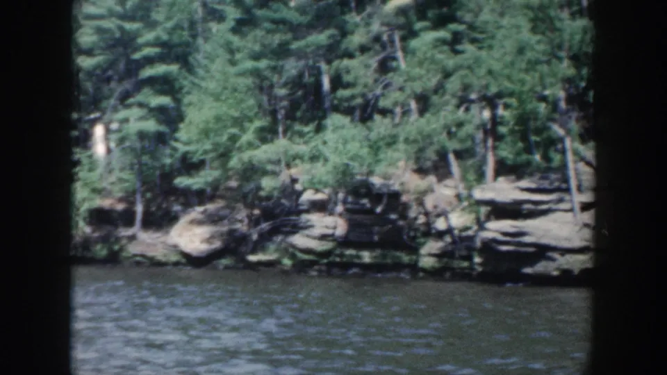 1964 Wisconsin 8mm frame with a dark window divider, textured lake water, and trees over layered shoreline rocks.