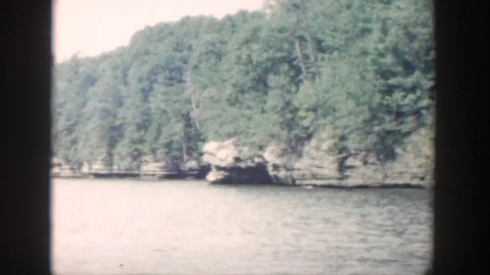 1964 Wisconsin 8mm view opening to a broader lake scene with wooded hills, distant rocks, and hazy exposure.
