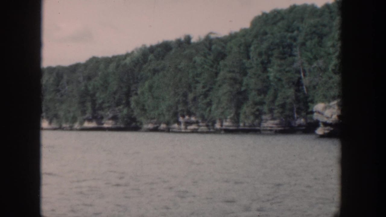 1964 Wisconsin 8mm lake view with dense trees, low rocky shore, gray water, and dark vertical framing from the filmed vantage point.