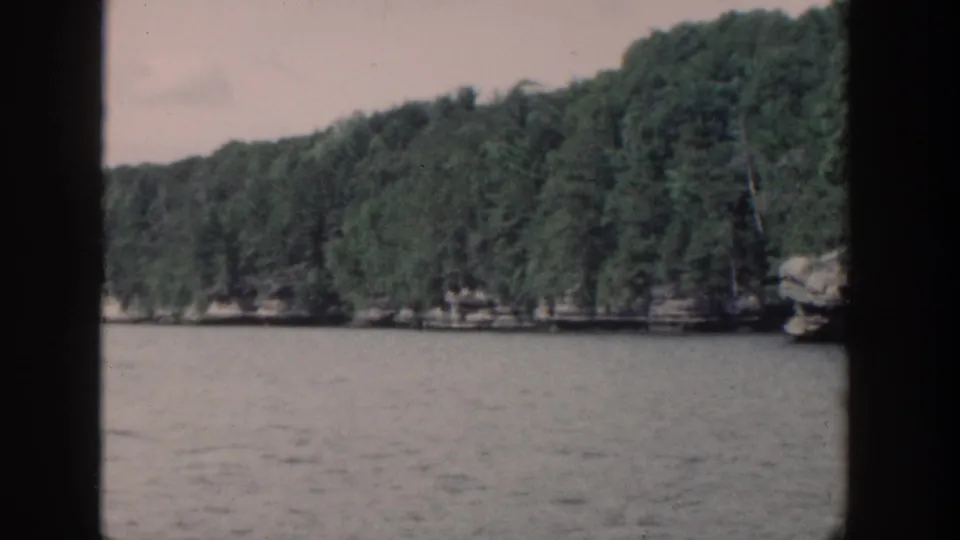 1964 Wisconsin 8mm lake view with dense trees, low rocky shore, gray water, and dark vertical framing from the filmed vantage point.