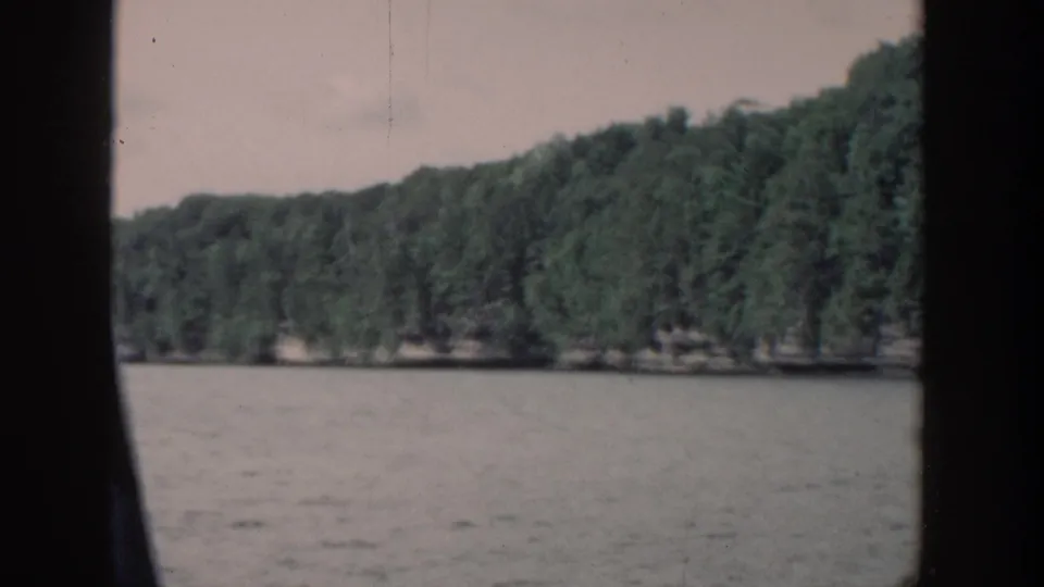 1964 Wisconsin home-movie frame showing water in the foreground and a continuous green shoreline in the distance, with dark side framing.