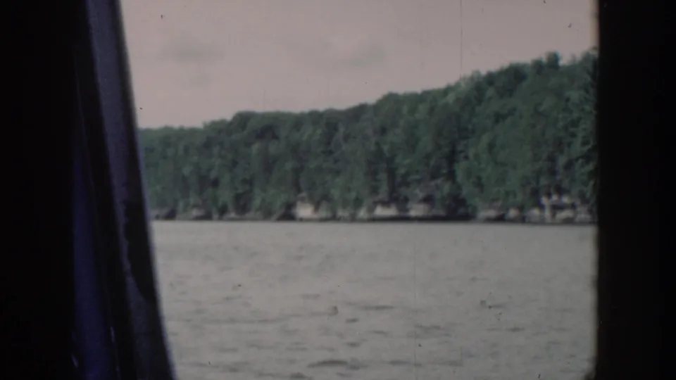 1964 Wisconsin 8mm frame of gray lake water and wooded shoreline, partly divided by a dark vertical boat-window or viewfinder edge.