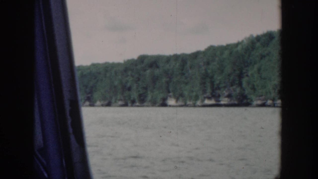 1964 Wisconsin lake shoreline viewed from inside a moving boat, with trees, water, pale sky, and dark window framing.