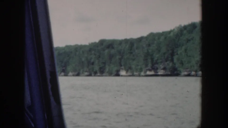 1964 Wisconsin lake shoreline viewed from inside a moving boat, with trees, water, pale sky, and dark window framing.