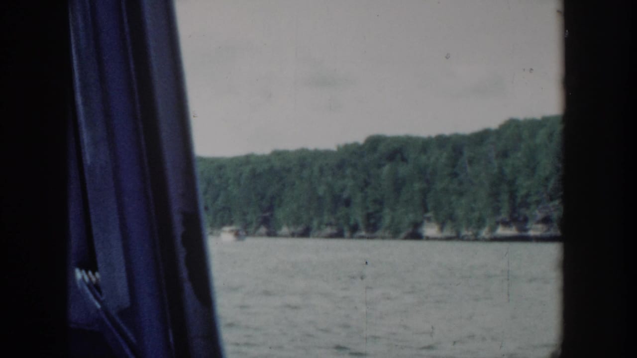 A passenger view across a Wisconsin lake shows distant trees and water through dark boat-window framing in 1964 home-movie footage.