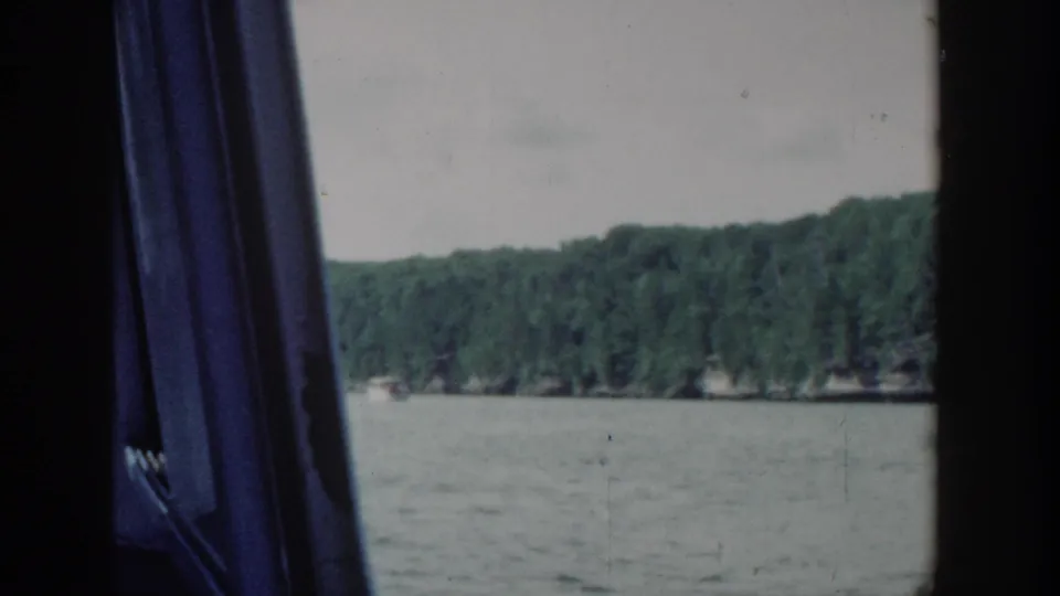 A passenger view across a Wisconsin lake shows distant trees and water through dark boat-window framing in 1964 home-movie footage.
