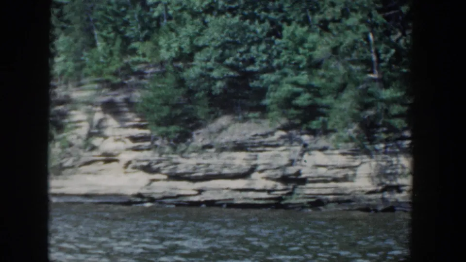Tree-covered rocky cliff and sedimentary ledges line the Wisconsin lakeshore in this 1964 8mm boat-view frame.