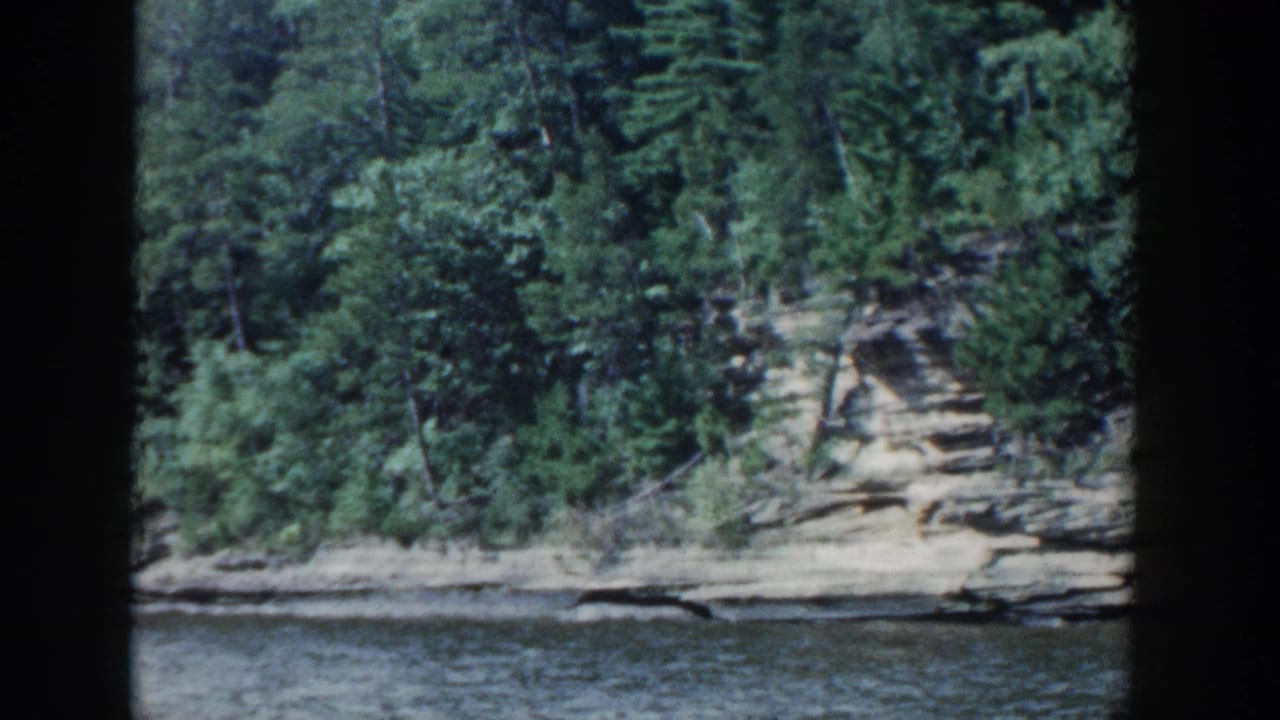 1964 Wisconsin 8mm view of calm lake water facing a rocky, tree-lined shoreline.