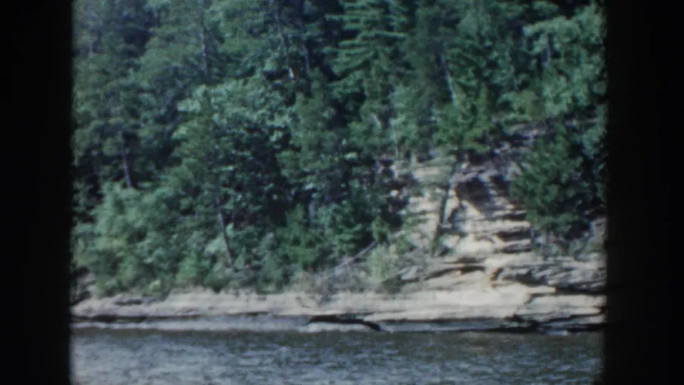 1964 Wisconsin 8mm view of calm lake water facing a rocky, tree-lined shoreline.