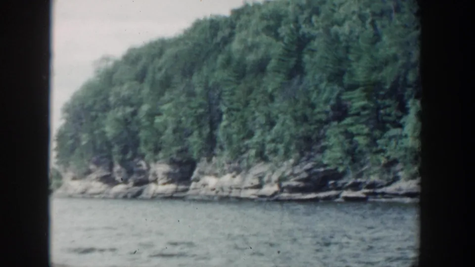 1964 Wisconsin lakefront view with forested shoreline, exposed rock, and calm water.