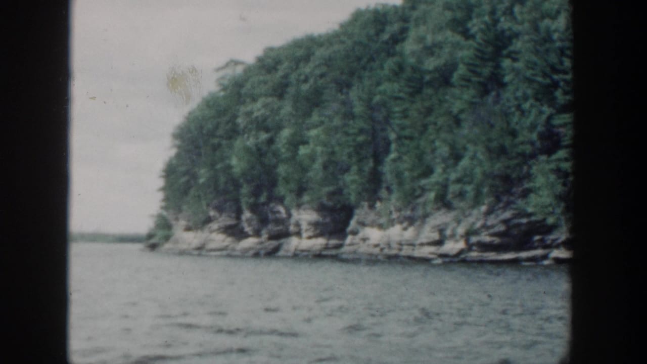 1964 Wisconsin 8mm frame showing a wooded rocky shoreline across lake water, filmed from a moving boat with dark frame edges.