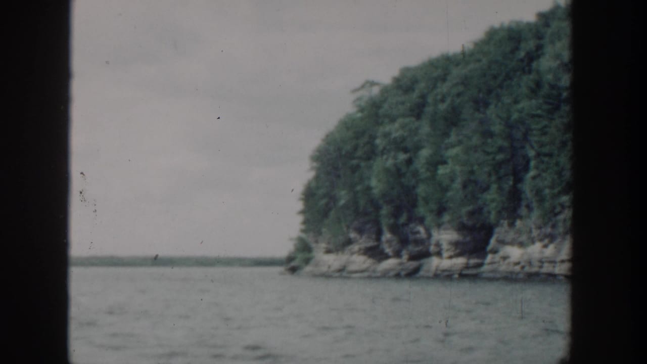 1964 Wisconsin home-movie frame with a wooded rocky point and lake horizon, viewed from a boat through dark film borders.