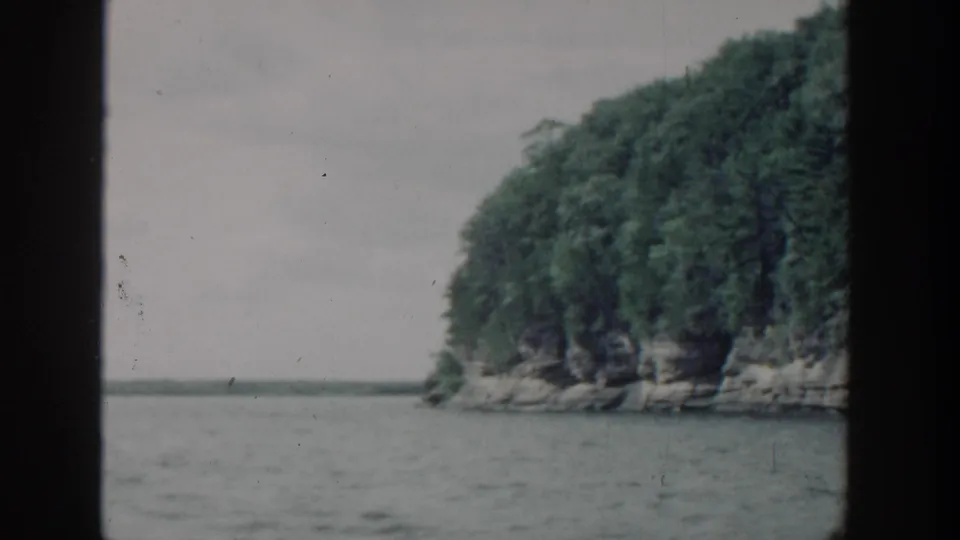 1964 Wisconsin home-movie frame with a wooded rocky point and lake horizon, viewed from a boat through dark film borders.