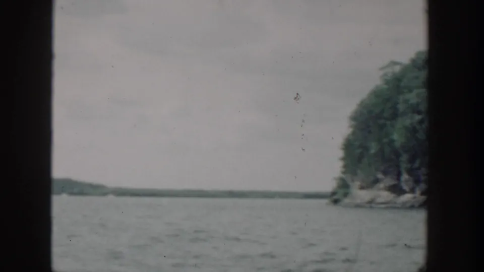 1964 Wisconsin 8mm frame showing lake water, a partial wooded shoreline, and low distant land under overcast light.