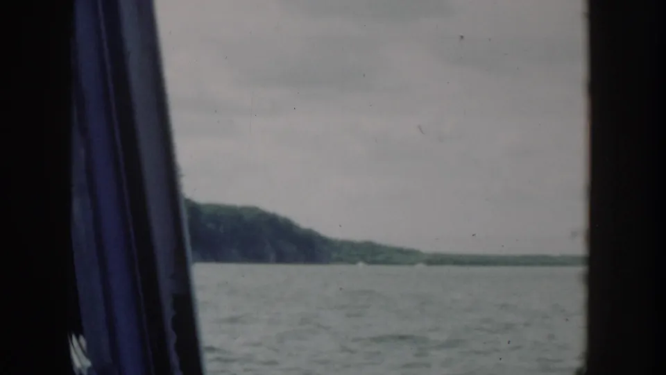 1964 Wisconsin 8mm frame showing calm river water and a distant wooded bank through a dark boat or window opening.
