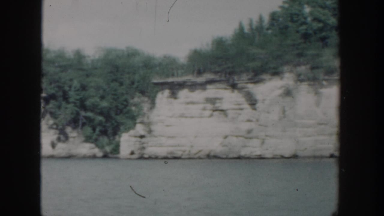 1964 Wisconsin 8mm frame of a white riverbank cliff topped with green trees, viewed from across the water.