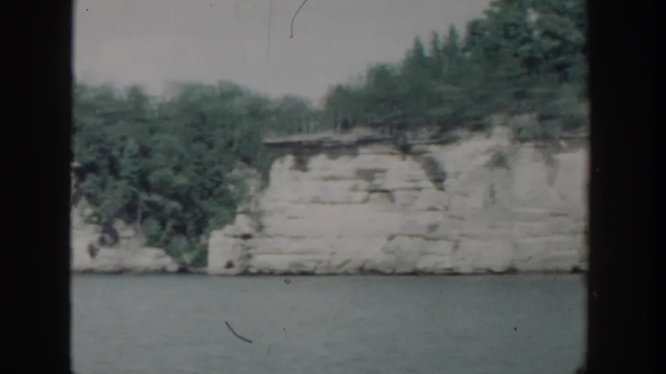 1964 Wisconsin 8mm frame of a white riverbank cliff topped with green trees, viewed from across the water.