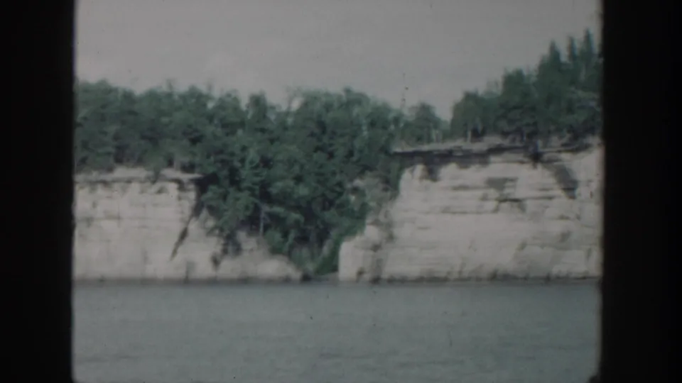 1964 Wisconsin 8mm frame showing a wooded white riverbank passing beside calm water, with dark framing at both sides.