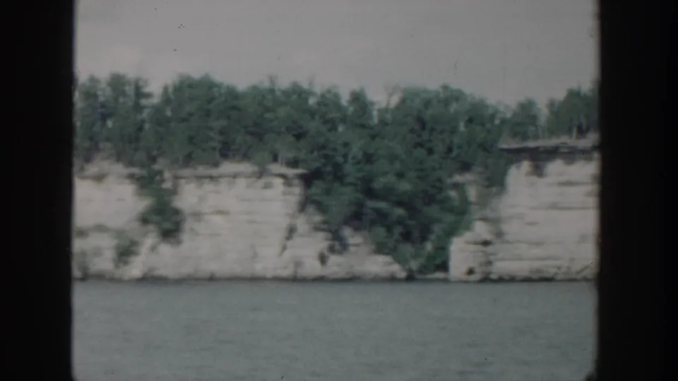 1964 Wisconsin 8mm frame of a white cliff shoreline and dense green trees, viewed from a moving river vantage point.
