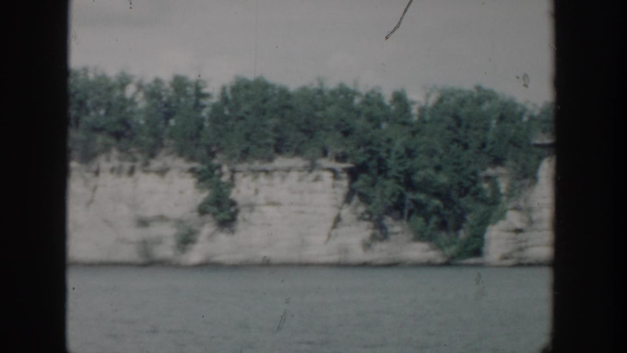 1964 Wisconsin 8mm frame showing a pale lakeside rock bluff topped with trees across calm water.