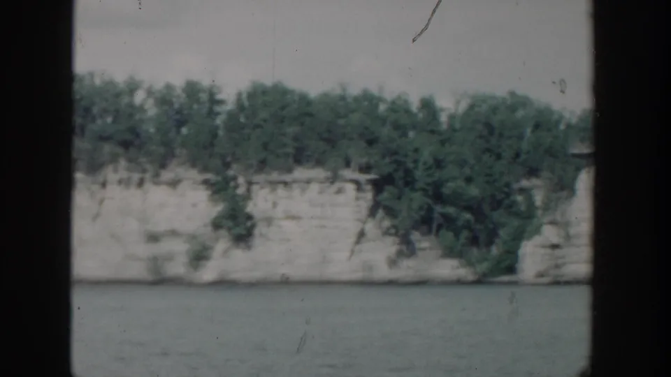 1964 Wisconsin 8mm frame showing a pale lakeside rock bluff topped with trees across calm water.