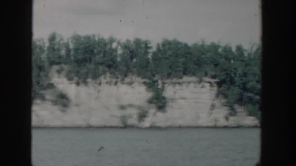 1964 Wisconsin home-movie view of a pale shoreline bluff, dense trees, and calm water.