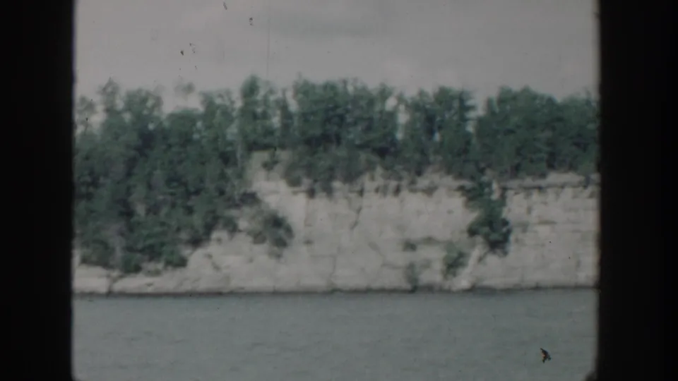 1964 Wisconsin 8mm frame with calm water in front of a pale bluff and thick green shoreline trees.