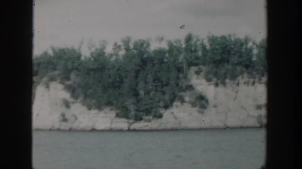 1964 Wisconsin home-movie frame showing a wooded pale bluff across water with visible film border and dust.