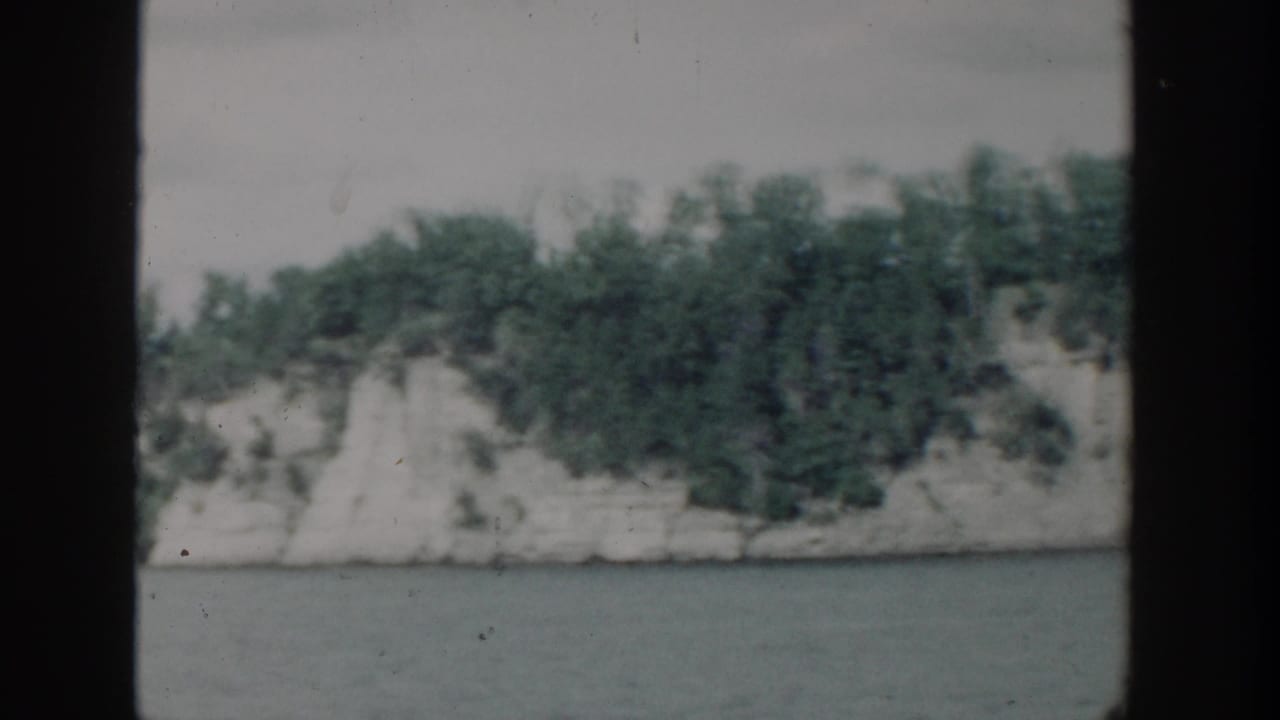 1964 Wisconsin 8mm frame showing a lake below a pale wooded cliff, seen in soft focus through film gate edges.