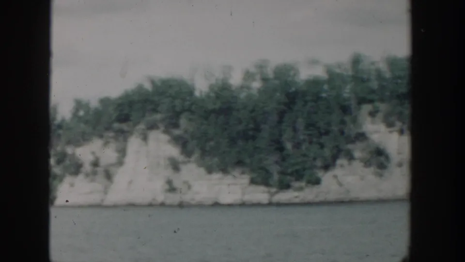 1964 Wisconsin 8mm frame showing a lake below a pale wooded cliff, seen in soft focus through film gate edges.