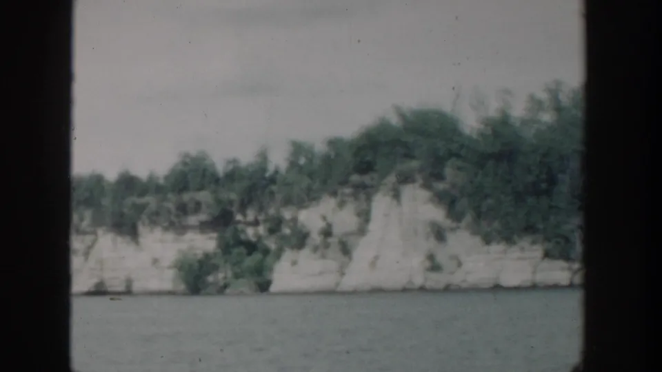 1964 Wisconsin home-movie view of a pale rocky shoreline and trees across still water, framed by dark 8mm scan borders.