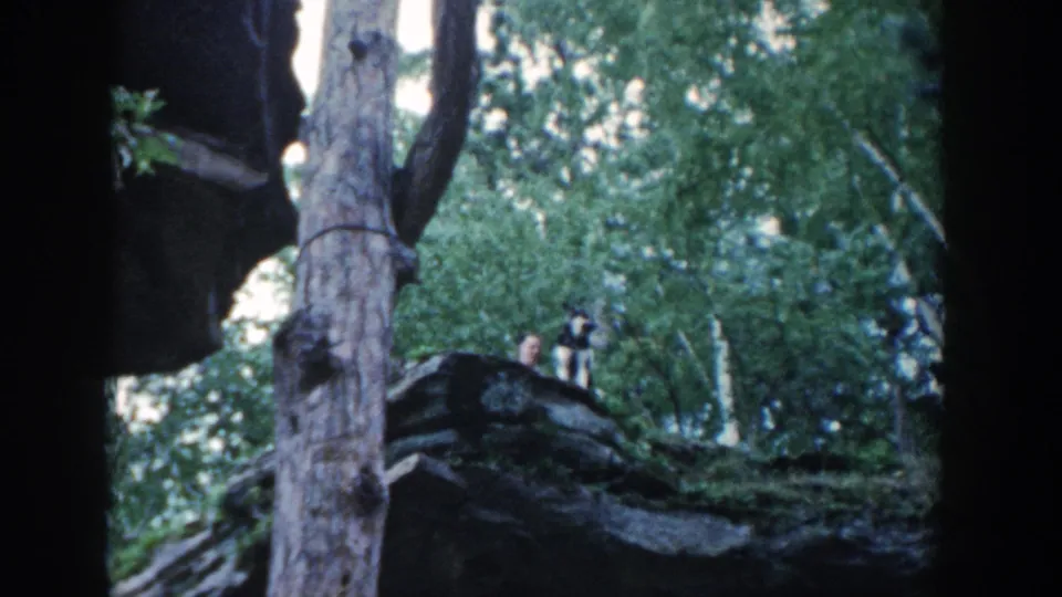 1964 Wisconsin 8mm frame of two distant people standing on a rocky wooded outcrop, with one appearing to hold a camera.